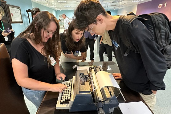 Três jovens ao redor de uma máquina de escrever, que está em cima de uma mesa de madeira. Menina à esquerda escreve na máquina de escrever, menina ao centro da imagem e menino à direita observam   