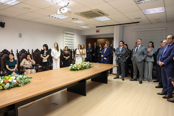 Fotografia horizontal: autoridades e convidados em  sala institucional com mesa de madeira e arranjos florais, cerimônia formal, grupo em pé observa solenidade, em tons bege, preto e verde