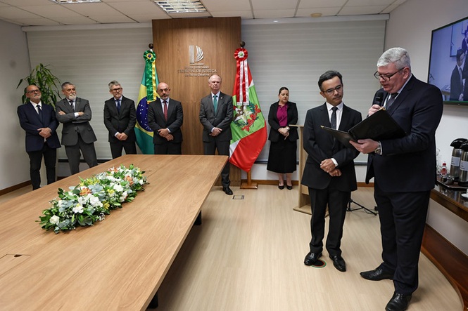 Fotografia horizontal: autoridades em cerimônia, sala institucional com mesa de madeira, ato formal, o desembargador Rubens Schulz e o Juíz André Luiz Dacol ao centro entre as bandeiras do Brasil e de Santa Catarina