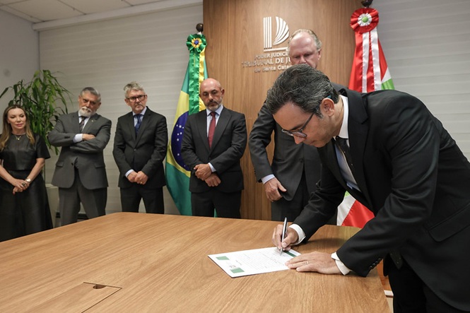 Fotografia horizontal: Magistrado assinando documento em sala institucional com mesa de madeira e bandeiras do Brasil e de Santa Catarina, em cerimônia formal enquanto outras autoridades observam