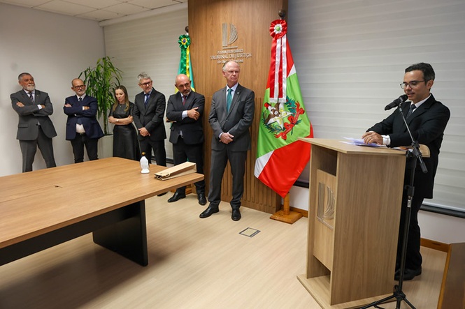 Fotografia horizontal: Humberto Goulart da Silveira discursa ao microfone enquanto o Desembargador Rubens Schulz e demais autoridades observam, em tons bege, verde e preto
