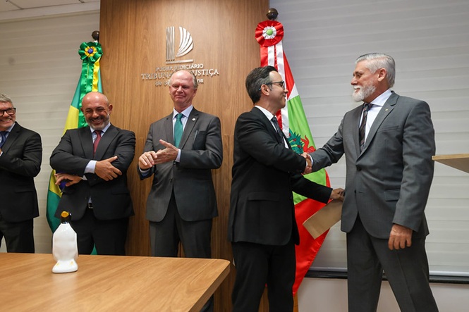 Fotografia horizontal: autoridades cumprimentando-se em sala institucional com bandeiras do Brasil e de Santa Catarina, em cerimônia formal 
