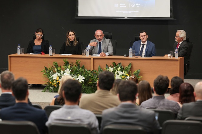 Foto colorida: mesa de debatedores diante do público, auditório institucional interno, evento formativo, conduzindo debate em bege, preto e verde