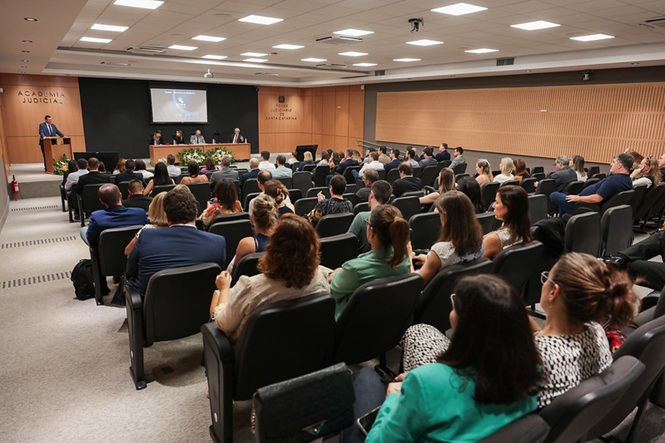 Foto colorida: público sentado em auditório amplo, Academia Judicial, evento institucional, acompanhando debate em bege, preto e verde