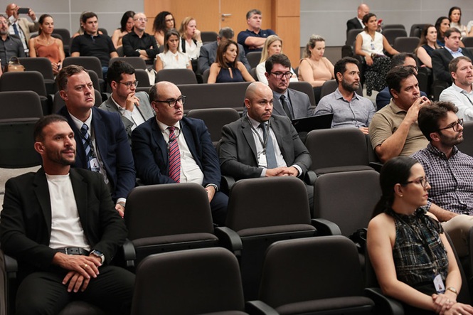 Foto colorida: público sentado em fileiras, auditório institucional interno, evento formativo, acompanhando apresentação em bege, preto e cinza