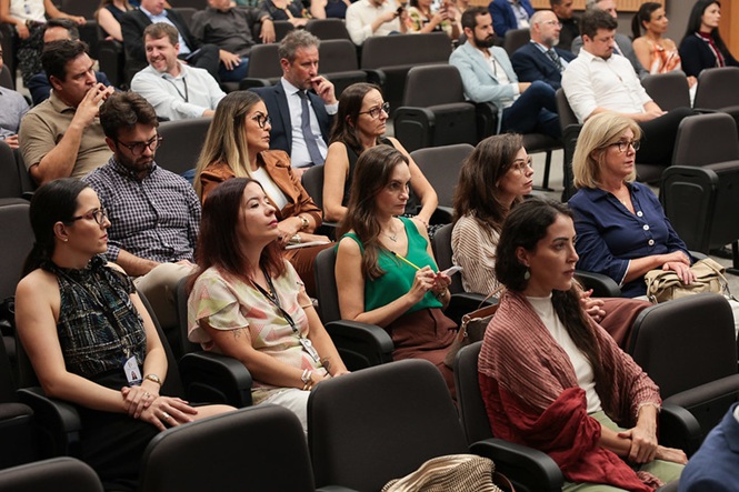 Foto colorida: público atento e fazendo anotações em auditório institucional, ambiente interno amplo, evento formativo, acompanhando palestra em bege, preto e cinza