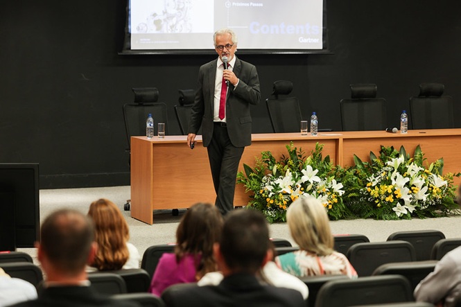 Foto colorida: palestrante com microfone diante do público, auditório institucional interno, evento formativo, apresentando conteúdo em preto, bege e verde