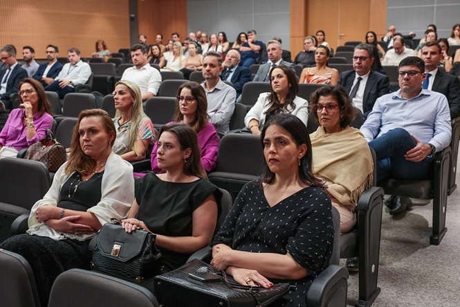 Foto colorida: público sentado em auditório institucional amplo, ambiente interno, evento formativo, acompanhando apresentação em bege, preto e cinza