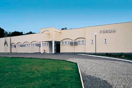 A imagem mostra a fachada de um fórum com uma arquitetura simples e horizontal, pintada em um tom de creme claro. O edifício é de um andar e possui uma série de janelas em arco, criando um estilo uniforme e ordenado. A entrada principal está levemente recuada, com uma estrutura de concreto que forma um pequeno pórtico decorativo. À frente do prédio, há um amplo gramado bem cuidado e um caminho pavimentado que leva até a entrada, com iluminação decorativa em postes brancos. O céu azul claro ao fundo realça a simplicidade e serenidade do ambiente, enquanto algumas árvores são visíveis ao longe.
