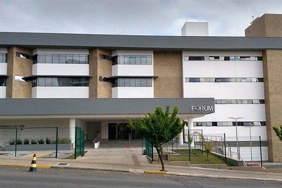 A imagem mostra a fachada de um edifício moderno, identificado como um fórum. O prédio tem linhas retas e uma aparência limpa, com cores neutras, principalmente branco e cinza. Há janelas amplas e retangulares distribuídas em três andares. Na entrada, há um pequeno pátio com calçada e uma rampa de acessibilidade, cercado por uma cerca baixa de metal verde. Um estacionamento está visível na frente do edifício, com um cone de trânsito próximo. O letreiro "FÓRUM" está em destaque na parte frontal direita do prédio. O ambiente ao redor tem vegetação e parece calmo.