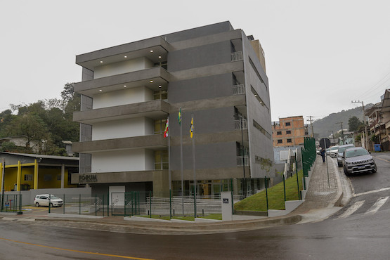 A imagem mostra um edifício moderno identificado como "FÓRUM," situado em uma rua inclinada. O prédio tem uma fachada cinza com varandas e é cercado por um muro baixo e grades metálicas. Na frente do edifício, há três mastros com bandeiras, sugerindo um ambiente institucional ou governamental. O dia está nublado, e a área ao redor parece urbana, com outras construções visíveis e uma estrada molhada, possivelmente devido à chuva. O local transmite um ar de formalidade e funcionalidade.
