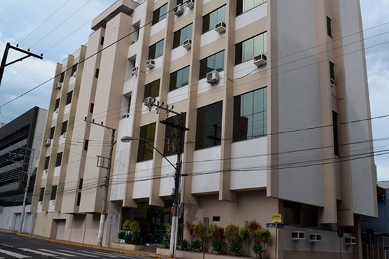 A imagem mostra um prédio institucional de vários andares, identificado como parte de uma comarca, com uma arquitetura funcional e janelas amplas, nas quais se encontram diversos aparelhos de ar-condicionado instalados. O edifício é de cor clara, com detalhes em branco e bege, e está localizado em uma rua urbana com postes de luz e fios elétricos visíveis. A vegetação paisagística na entrada inclui pequenos arbustos, e a fachada sugere que o prédio serve propósitos administrativos ou judiciais.