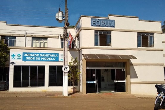 A imagem mostra a fachada de um edifício institucional com a palavra "FÓRUM" destacada na parte superior. Ao lado, há uma outra seção do prédio identificada como "UNIDADE SANITÁRIA SEDE DE MODELO," que pertence a um serviço de saúde. O prédio tem uma arquitetura simples e funcional, com paredes claras e janelas grandes. Na frente, há uma cobertura de vidro e um piso de concreto, indicando uma entrada principal. O ambiente parece ser uma área central de uma cidade, com calçadas e postes de iluminação pública.