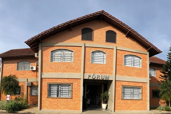 A imagem mostra a fachada de um edifício identificado como "FÓRUM," com uma construção em tijolo aparente e detalhes em concreto. O prédio tem um telhado inclinado com telhas marrons escuras e janelas com grades de metal. A arquitetura é simples e funcional, com um toque rústico devido aos tijolos expostos. Há uma entrada central destacada, com vasos de plantas decorando o local, e o entorno possui vegetação, como árvores e arbustos, que complementam o ambiente de forma agradável.