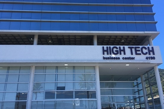 A imagem mostra a fachada de um edifício moderno identificado como "HIGH TECH business center," com o número 4190 exibido ao lado do nome. A estrutura é composta por vidro e concreto, destacando um design contemporâneo. O prédio tem grandes janelas de vidro que refletem o céu e parte do ambiente ao redor, e há um amplo espaço coberto na entrada. A arquitetura e o nome indicam que o local provavelmente abriga escritórios ou espaços corporativos de alta tecnologia.