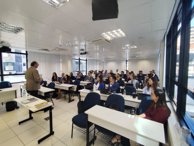 fotografia mostra uma sala de aula ampla e iluminada, com magistrados sentados em fileiras de cadeiras azuis com mesas brancas, acompanhando uma exposição. À frente da sala, o desembargador Volnei Carlin de pé fala ao grupo, posicionado próximo a uma mesa com materiais e equipamentos. O ambiente possui grandes janelas laterais de vidro, teto com iluminação embutida e projetor, compondo um espaço institucional de formação e aprendizado.