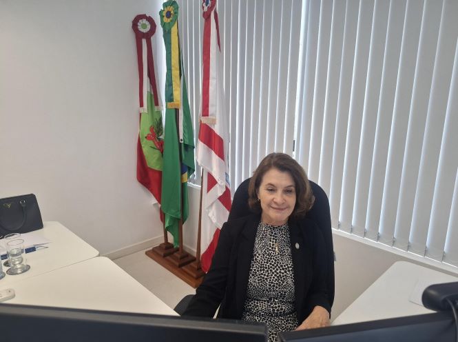 Desembargadora Vera Lúcia Ferreira Copetti sentada em uma cadeira na frente de dois computadores, ao fundo as bandeiras do Brasil, Santa Catarina e do Poder Judiciário