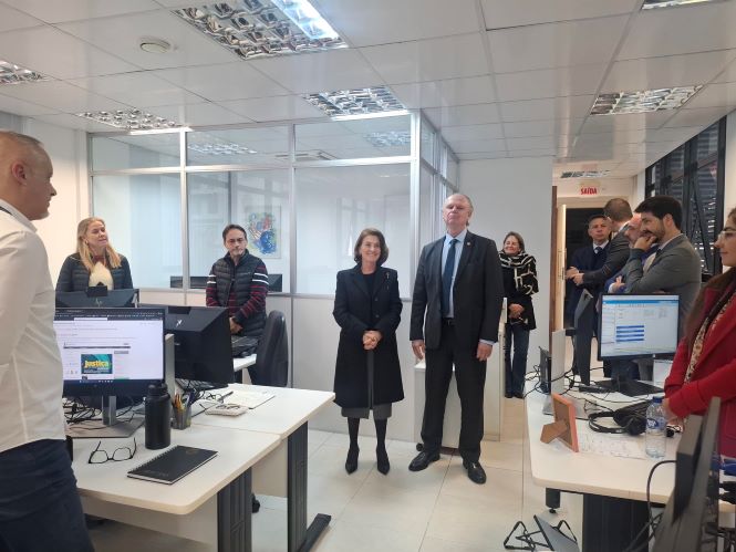 Fotografia mostra uma visita institucional em ambiente de trabalho do Judiciário. Ao centro da imagem estão a desembargadora Vera Lúcia Copetti e o presidente do TJSC, desembargador Rubens Schulz, em pé, lado a lado, enquanto observam o espaço. Ao redor, servidores acompanham a visita próximos às estações de trabalho com computadores, em sala ampla e iluminada.