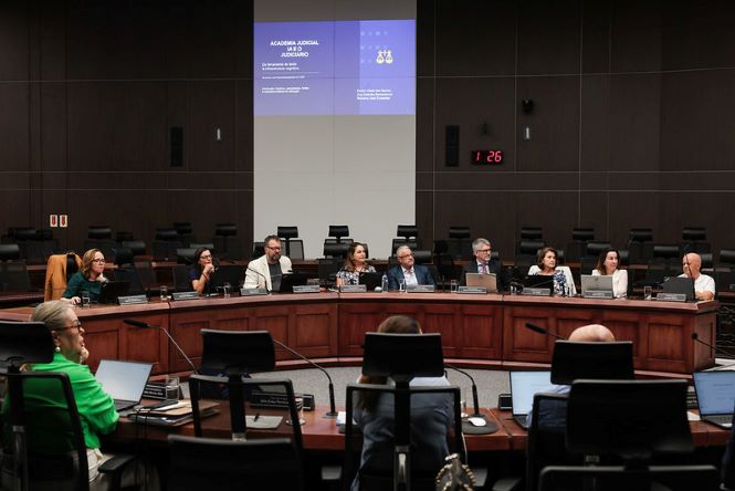 Imagem mostra parte do plenário do Tribunal de Justiça de Santa Catarina durante reunião institucional. Ao centro, magistrados e servidores estão sentados lado a lado em uma bancada semicircular de madeira escura, olhando para a direita, em direção a uma apresentação. Em primeiro plano, aparecem cadeiras e mesas com computadores voltados para o centro do plenário. Ao fundo, um telão projeta a identidade visual da apresentação.