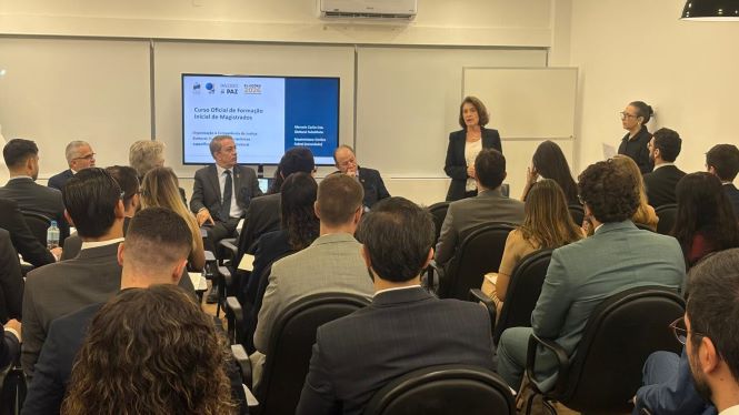 Em uma sala de aula, a desembargadora Vera Lúcia Ferreira Copetti está em pé à frente de um grupo de magistrados em formação, dirigindo-se aos participantes do curso. Ao lado dela, três desembargadores estão sentados em poltronas, compondo a mesa de abertura. Os alunos, vestidos com trajes formais, ocupam cadeiras organizadas em fileiras e estão voltados para a palestrante. Ao fundo, um monitor exibe a apresentação do “Curso Oficial de Formação Inicial de Magistrados”, com identidade visual institucional.