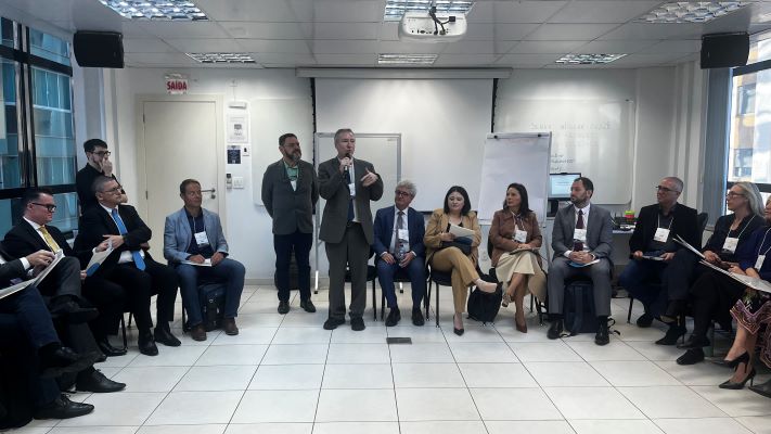 Sala interna de ambiente corporativo, com piso claro em cerâmica, paredes brancas e iluminação artificial por luminárias retangulares no teto. Ao fundo, há um quadro branco com anotações pouco legíveis, um flip chart e um projetor fixado no teto. Uma porta à esquerda exibe uma placa vermelha com a palavra “SAÍDA”. As cadeiras estão organizadas em semicírculo, ocupadas por cerca de 12 a 15 pessoas vestidas com roupas formais, como ternos e conjuntos sociais. No centro do grupo, o juiz federal Vladimir Vitovsky, em pé, segura um microfone e parece conduzir a fala. Ao lado, Erisevelton Lima permanece em pé, próxima à parede. As demais pessoas estão sentadas, algumas segurando papéis ou cadernos.