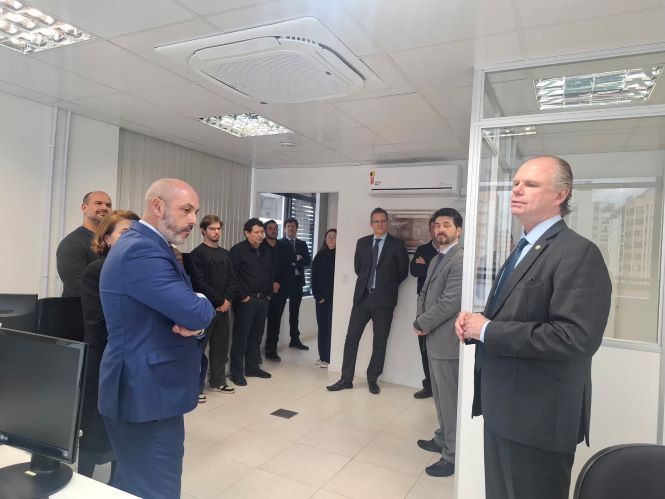 Fotografia mostra o presidente do TJSC, desembargador Rubens Schulz, à direita da imagem, falando a um grupo de magistrados, servidores e colaboradores reunidos em uma sala administrativa. Os participantes acompanham a fala em pé, próximos às estações de trabalho e computadores, em ambiente interno iluminado.