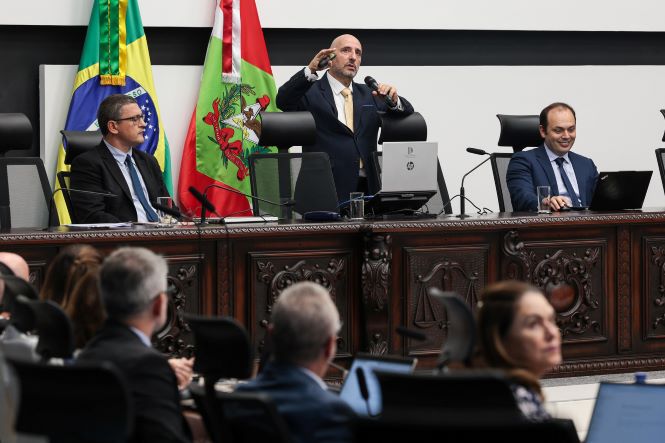 Imagem registra uma reunião institucional no plenário do Tribunal de Justiça de Santa Catarina. Ao centro, um homem de terno escuro e gravata clara, o juiz Romano Enzweiler, está em pé atrás da bancada principal, segurando um microfone e gesticulando com uma das mãos enquanto fala ao público. Ao seu lado, dois homens também de terno estão sentados em cadeiras altas atrás da mesa de madeira escura entalhada; um observa atentamente e o outro sorri diante de um computador aberto. Ao fundo, estão posicionadas as bandeiras do Brasil e do estado de Santa Catarina. Em primeiro plano, aparecem desfocados participantes sentados de frente para a mesa principal, compondo o ambiente formal e solene do plenário.
