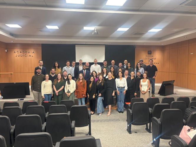 Fotografia em ambiente interno mostra um grupo grande de aproximadamente 30 pessoas posando para uma foto em um auditório. Elas estão organizadas em duas fileiras, a maioria em pé, sorrindo e voltada para a câmera. O espaço possui cadeiras pretas dispostas em fileiras no primeiro plano, parcialmente vazias. Ao fundo, há um palco com uma tela de projeção central e paredes revestidas em madeira clara. À esquerda, lê-se “Academia Judicial” na parede; à direita, há o brasão institucional e a inscrição “Poder Judiciário de Santa Catarina”. A iluminação é clara e uniforme, vinda do teto. As pessoas vestem roupas variadas, em estilo casual e profissional.