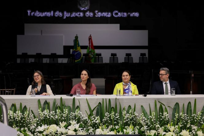 Imagem de uma mesa de evento em um ambiente institucional amplo, com palco ao fundo. Na parte superior, há a inscrição iluminada “Tribunal de Justiça de Santa Catarina”. Atrás da mesa, duas bandeiras estão posicionadas ao centro, e cadeiras vazias compõem o cenário do palco. Em primeiro plano, quatro pessoas estão sentadas lado a lado atrás de uma mesa longa coberta por toalha clara. Sobre a mesa, há copos de vidro com água, microfones e papéis. A base da mesa é decorada com arranjos de flores brancas e folhagens verdes. Da esquerda para a direita, conforme informado no contexto: Haydee Lopnow, Alessandra Ludvig, desembargadora Haidee Meneguzzi e o professor Vanderlei de Oliveira Farias. Cada pessoa está voltada para a frente, em posição de participação em mesa de debate ou palestra. A iluminação é direcionada para a mesa, destacando as pessoas em primeiro plano, enquanto o fundo permanece mais escuro e desfocado, reforçando o caráter formal e institucional do evento.