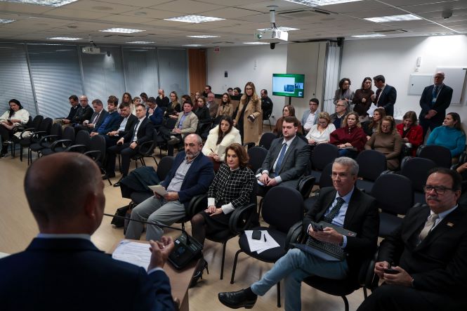 Fotografia de uma sala de eventos com dezenas de participantes sentados em cadeiras organizadas em fileiras. Em primeiro plano, aparece parcialmente a pessoa que conduz a atividade, vista de costas, falando ao público. Os participantes acompanham a apresentação, alguns com documentos ou dispositivos eletrônicos nas mãos. Ao fundo, há pessoas em pé, um monitor com conteúdo exibido e paredes envidraçadas com persianas. O ambiente é iluminado por luzes embutidas no teto.