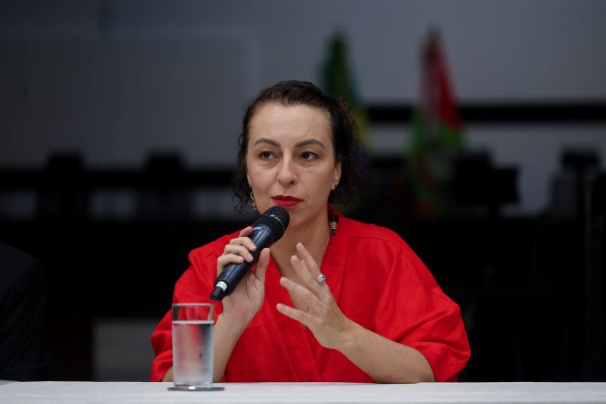Fotografia em plano aproximado mostra Carolina Rodrigues Costa sentada à mesa durante o evento. Ela veste blusa vermelha e segura um microfone com a mão direita enquanto fala. Com a mão esquerda, faz um gesto durante sua manifestação. À sua frente há um copo com água sobre a mesa. Ao fundo, desfocado, aparecem parte do auditório e das bandeiras posicionadas atrás da mesa de autoridades.