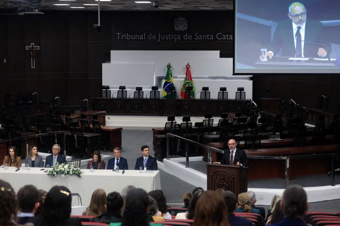 Auditório do Tribunal de Justiça de Santa Catarina visto de frente. Em primeiro plano, participantes acompanham a cerimônia sentados na plateia. À direita, André Luiz Dacol, presidente do TJSC em exercício, está no púlpito realizando um pronunciamento. A imagem de André Luiz Dacol também é exibida em destaque no telão instalado na parte superior do plenário. À esquerda, autoridades compõem a mesa principal, decorada com arranjo de flores. Ao fundo, estão as bandeiras do Brasil e de Santa Catarina e a estrutura do plenário do Tribunal.