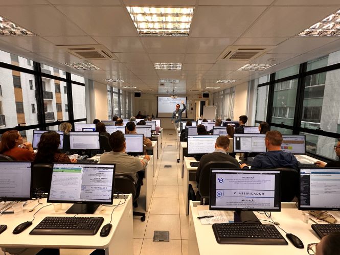 Fotografia de uma sala de informática durante uma atividade de capacitação. Participantes estão sentados em fileiras, cada um utilizando um computador de mesa. Ao centro da sala, há um corredor que leva até a área do instrutor, posicionada em frente a uma tela de projeção. Nos monitores visíveis, aparecem conteúdos relacionados ao sistema eproc e a ferramentas de automação. O ambiente é iluminado por luzes no teto, possui grandes janelas nas laterais e vista para edifícios vizinhos. As mesas e paredes são claras, e a disposição dos equipamentos evidencia uma atividade de treinamento presencial com diversos participantes.