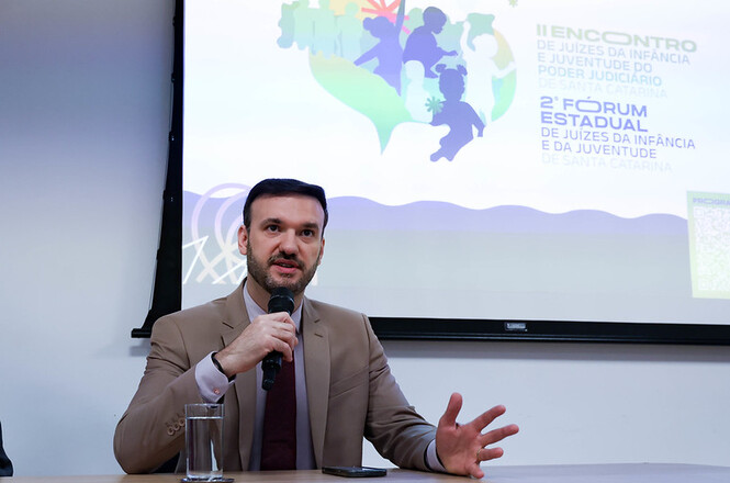 Na imagem juiz Fernando Machado Carboni fala ao microfone, ao fundo um painel com a identidade visual do evento
