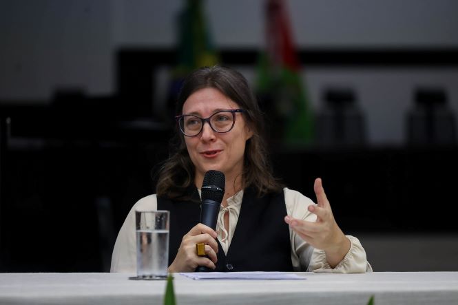 Fernanda Loppnow está sentada atrás de uma mesa, em um ambiente interno que lembra um auditório ou sala de eventos. Ela segura um microfone com uma das mãos enquanto faz um gesto com a outra, como se estivesse explicando ou falando ao público. Sobre a mesa, à frente, há um copo transparente com água. A pessoa usa óculos de armação escura, veste uma camisa clara com mangas compridas e um colete escuro por cima. Ao fundo, há bandeiras desfocadas e móveis escuros, sugerindo um ambiente institucional. A iluminação é suave, com foco principal na pessoa em primeiro plano.