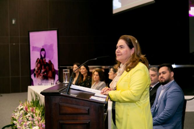 Fotografia mostra a desembargadora Hildemar Meneguzzi de Carvalho, coordenadora da Cevid, em pé atrás de um púlpito de madeira, durante um evento. Ela veste um blazer amarelo-claro e fala ao público, diante de um microfone. Sobre o púlpito há documentos e um segundo microfone. Ao fundo, outras pessoas estão sentadas em uma mesa, acompanhando a atividade. À esquerda, há um painel com uma imagem de divulgação do evento e, na parte inferior, um arranjo de flores. O ambiente é um auditório, com paredes escuras e iluminação direcionada ao palco.
