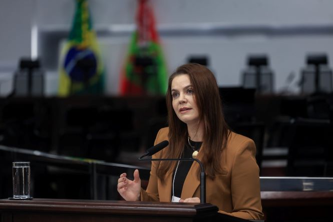 Fotografia horizontal de Juliana Motim de Oliveira, em pé diante de um púlpito de madeira, durante seu pronunciamento. Ela tem cabelos longos castanhos e veste blazer marrom sobre blusa preta, com um cordão de identificação no pescoço. Juliana gesticula com uma das mãos enquanto fala ao microfone. Ao fundo, aparecem as bandeiras do Brasil e de Santa Catarina, além de elementos do plenário. Há um copo com água sobre o púlpito, à esquerda.