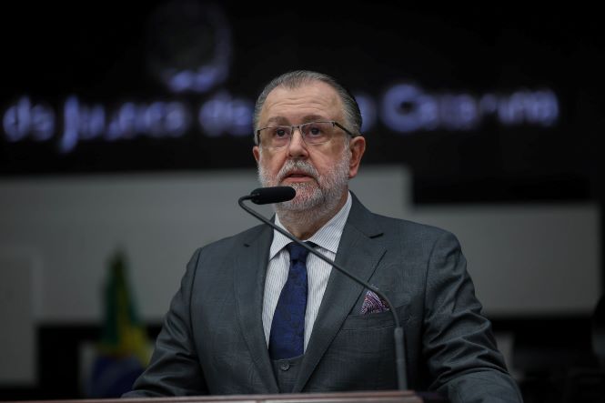 Pessoa identificada no contexto como desembargador Marcio Rocha Cardoso está sentada atrás de uma mesa em um ambiente que parece ser um auditório institucional. A pessoa segura um microfone próximo à boca, em posição de fala, voltada em direção ao público. Sobre a mesa há um copo transparente com água. A pessoa veste traje formal composto por terno escuro, camisa clara e gravata. Ao fundo, há um painel escuro com texto parcialmente visível e desfocado, além de bandeiras também desfocadas, sugerindo um ambiente oficial, possivelmente ligado ao sistema de justiça. A iluminação destaca a pessoa em primeiro plano, com o restante do ambiente em segundo plano menos nítido.