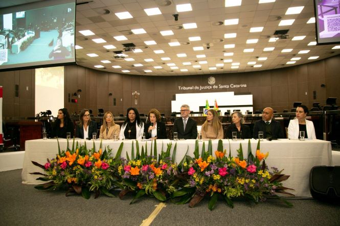 Fotografia colorida registrada no plenário do Tribunal de Justiça de Santa Catarina. Em primeiro plano, uma longa mesa coberta por toalha branca está posicionada diante de um grande arranjo floral com flores em tons de laranja, rosa, amarelo e roxo. Sentadas atrás da mesa, da esquerda para a direita, estão a juíza de direito Fernanda Pereira Nunes, de cabelos escuros e roupa preta; Naiara Brancher, de óculos e blazer bege; a juíza de direito Andréa Rezende Russo, de cabelos claros e blazer bege; a desembargadora Tânia Regina Silva Reckziegel, de cabelos escuros e traje preto; a desembargadora Vera Lúcia Copetti, que fala ao microfone, usando roupa clara; Dinart Francisco Machado, de terno escuro e gravata; Quézia de Araújo Duarte Nieves Gonzales, de cabelos claros e blazer bege; Eliana Volcato Nunes, de cabelos grisalhos e traje escuro; Mauro Candido, de terno escuro; e Carolina Rodrigues Costa, de blazer branco. Ao fundo, vê-se a bancada do plenário, as bandeiras do Brasil, de Santa Catarina e do Poder Judiciário, além da inscrição “Tribunal de Justiça de Santa Catarina” na parede. Monitores suspensos exibem imagens do evento.