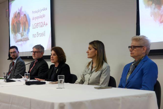 Fotografia mostra cinco participantes sentados à mesa de abertura do seminário Proteção Jurídica da População LGBTQIA+ no Brasil, coberta por uma toalha branca. Da esquerda para a direita estão: Túlio Parreiras, de terno cinza, camisa branca e gravata vermelha; Dinart Machado, de terno escuro, camisa preta e gravata vermelha; Vera Copetti, de blazer preto, falando ao microfone; Cristine Mattos, com blusa cinza-clara e cabelos lisos na altura dos ombros; e Margareth Hernandes, de blazer azul, camisa estampada e óculos de armação escura. À frente de cada participante há um copo com água e, ao fundo, dois telões exibem a identidade visual do evento.