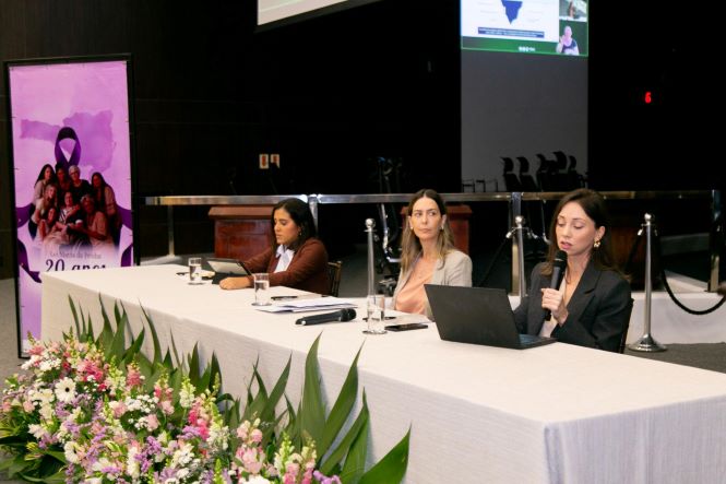 Fotografia mostra três mulheres sentadas atrás de uma mesa durante um evento. À direita, a defensora pública de Santa Catarina Anne Teive Auras segura um microfone e fala ao público, enquanto utiliza um notebook. Ao centro, está a defensora pública Luísa Rotondo Garcia, que acompanha a atividade. À esquerda, está a defensora pública de Santa Catarina Tainá Braga de Oliveira, sentada diante de um notebook. Sobre a mesa, coberta por uma toalha branca, há microfones, copos com água, documentos e equipamentos eletrônicos. Na parte inferior da imagem, há um grande arranjo de flores. Ao fundo, vê-se um painel com material de divulgação dos 20 anos da Lei Maria da Penha e uma tela de projeção.