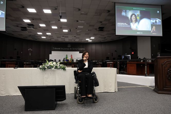 No plenário do Tribunal de Justiça de Santa Catarina, Flávia Cintra está sentada em uma cadeira de rodas à frente da mesa principal, segurando um microfone durante sua palestra. Sobre a mesa há um arranjo de flores brancas. Ao fundo, aparecem as bandeiras do Brasil e de Santa Catarina. Em um telão, é exibida a imagem da palestrante acompanhada do título de sua apresentação. O ambiente é amplo, com iluminação clara e estrutura de auditório.