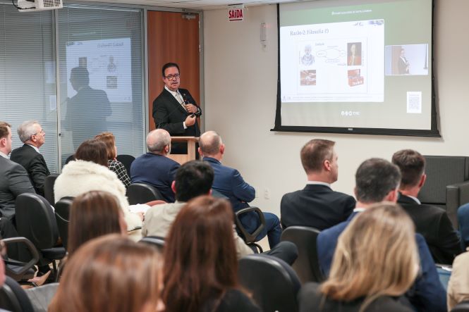 Fotografia de uma palestra em auditório. À frente da sala, Fernando de Lima Neto está em pé, ao lado de um púlpito, segurando um microfone e apresentando um conteúdo projetado em uma tela. O público, composto por dezenas de participantes sentados em cadeiras, acompanha a exposição voltado para a apresentação. O ambiente é uma sala de eventos corporativa, com iluminação clara e projetor exibindo slides ao fundo.