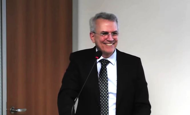 Alexandre Freire Pimentel está em pé junto a um púlpito, durante uma apresentação em ambiente interno. Veste terno escuro, camisa branca e gravata com detalhes claros. À sua frente há um microfone de mesa, e ao fundo aparecem uma parede clara e uma porta de madeira. A fotografia registra o momento da palestra em plano próximo.