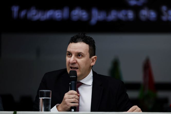Pessoa identificada no contexto como Alexsander Postali está sentada atrás de uma mesa em um ambiente institucional, semelhante a um auditório. A pessoa segura um microfone com uma das mãos e está em posição de fala voltada ao público. Sobre a mesa há um copo transparente com água. A pessoa veste traje formal, com terno escuro, camisa clara e gravata. Ao fundo, há um painel escuro com texto desfocado e bandeiras também desfocadas, indicando um ambiente oficial, possivelmente ligado ao sistema de justiça. A iluminação destaca a pessoa em primeiro plano.