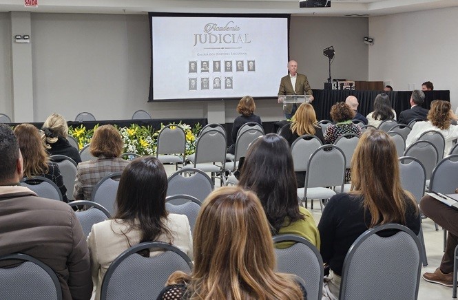 Copilot said: Ambiente de auditório com público sentado em cadeiras voltadas para o palco. Ao fundo, uma tela exibe “Academia Judicial” e “Galeria dos Diretores Executivos”. No centro, Rubens Schulz está no púlpito transparente, discursando. Há arranjos florais à frente do palco e equipamentos de iluminação ao lado.