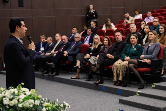 Em auditório com poltronas vermelhas, Rodrigo Lima está em pé à frente da plateia, segurando um microfone e dirigindo-se ao público. Nas primeiras fileiras, autoridades e convidados acompanham a apresentação sentados. Em primeiro plano, há um arranjo de flores brancas próximo ao palco. Ao fundo, mais participantes ocupam os assentos do auditório.