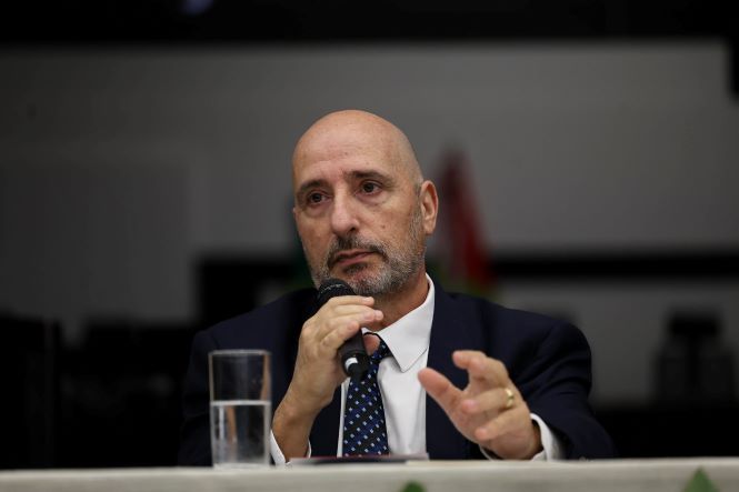 Fotografia em plano aproximado mostra o juiz Romano Enzweiler sentado à mesa durante o evento. Ele aparece usando terno escuro, camisa branca e gravata azul-marinho com pequenos pontos claros. Segurando um microfone com a mão esquerda, faz um gesto com a mão direita enquanto fala. À sua frente há um copo com água sobre a mesa. O fundo desfocado revela parte do auditório e das bandeiras posicionadas atrás da mesa de autoridades.