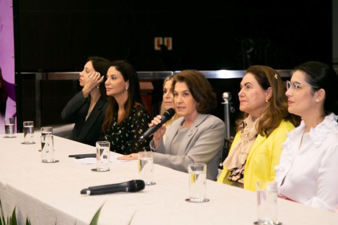 Fotografia mostra cinco mulheres sentadas lado a lado à mesa de um evento. Ao centro, a desembargadora Vera Lúcia Ferreira Copetti segura um microfone e fala ao público. À sua esquerda, estão duas mulheres; à direita, a desembargadora Hildemar Meneguzzi de Carvalho, coordenadora da Cevid, veste um blazer amarelo, seguida por outra mulher de camisa branca. Sobre a mesa, coberta por toalha branca, há copos com água e microfones. Ao fundo, vê-se um ambiente de auditório com iluminação reduzida.