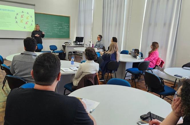 Foto horizontal: palestrante apresenta conteúdo em sala de aula para participantes sentados em mesas.